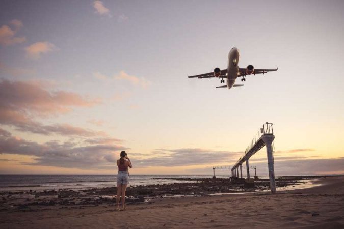 Une femme photographie un avion en vol bas au-dessus d'une plage au coucher du soleil, une scène inspirante pour voyager pas cher.