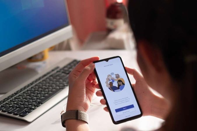Une personne tient un smartphone affichant la page d'accueil de l'application Microsoft Teams devant un ordinateur.