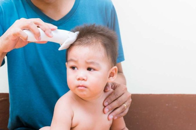 Un bébé au regard attentif se fait tondre les premiers cheveux fins par un adulte portant un t-shirt bleu.